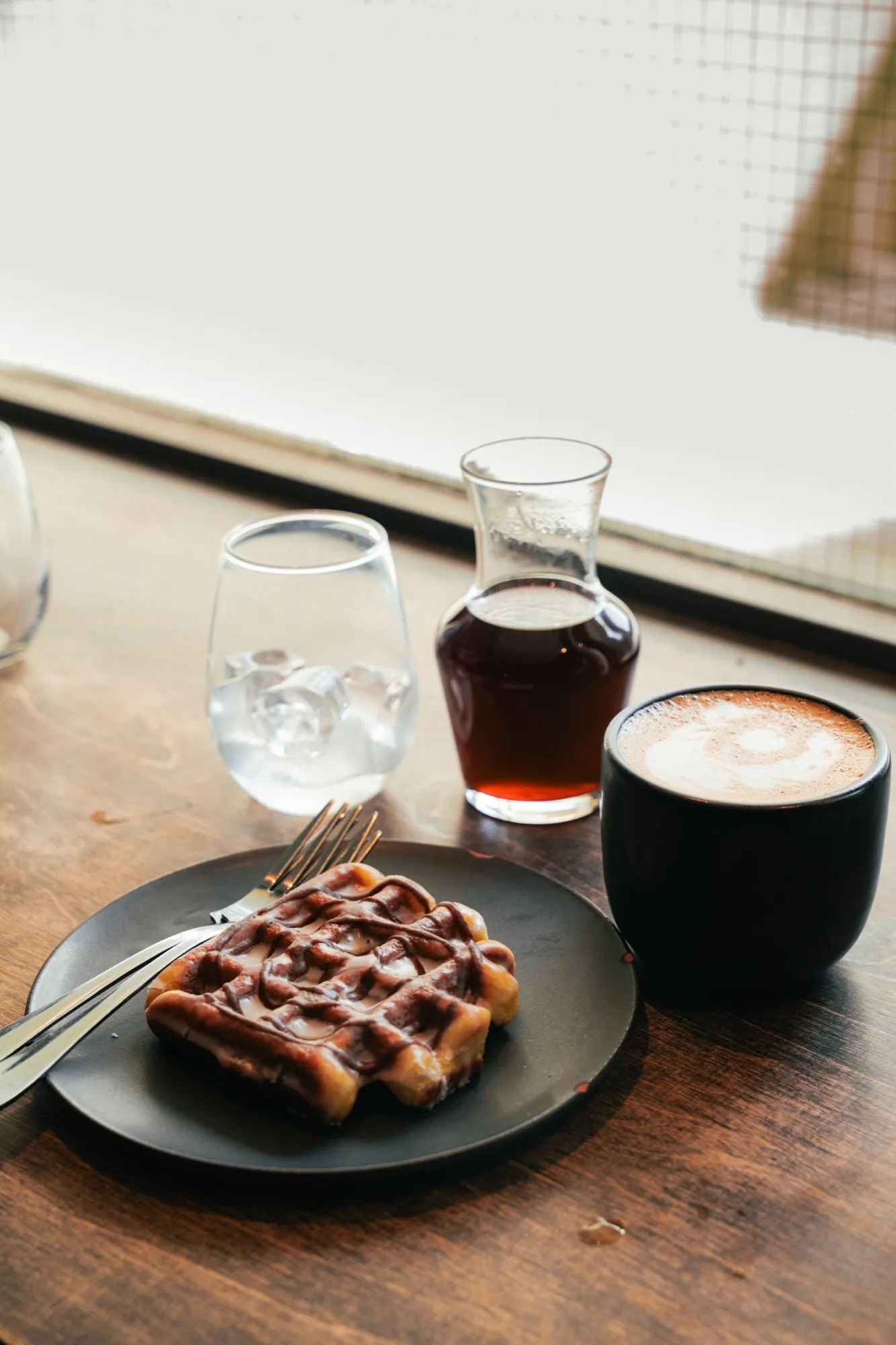 Chocolate drizzled waffle with latte art and iced coffee on wooden cafe table by window